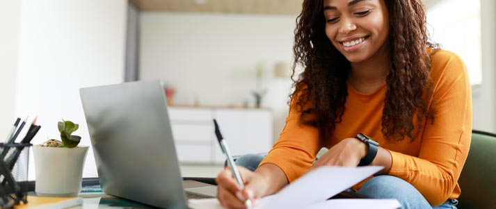 A young woman writing in a notebook while sat at her laptop