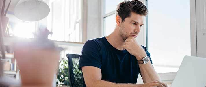 young man looking at laptop screen sat on chair by desk